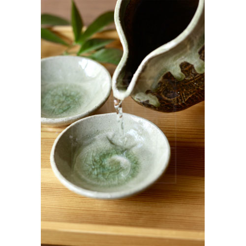 Green-glazed sake server and shallow cups with natural textures, styled with seasonal foliage.